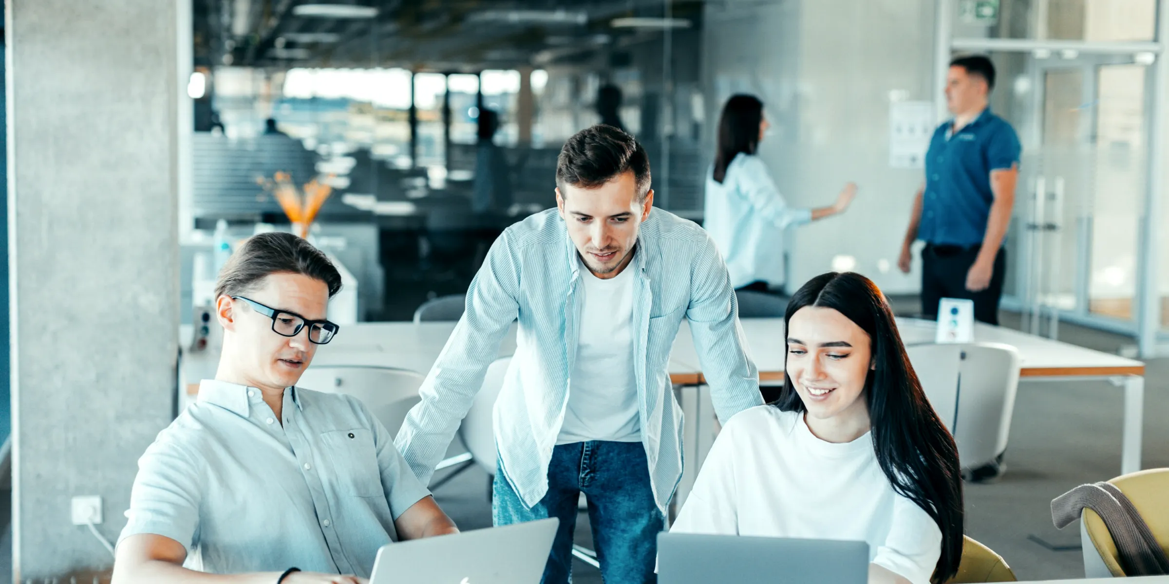 Un collega che si china ad aiutare due persone al computer in un ufficio luminoso