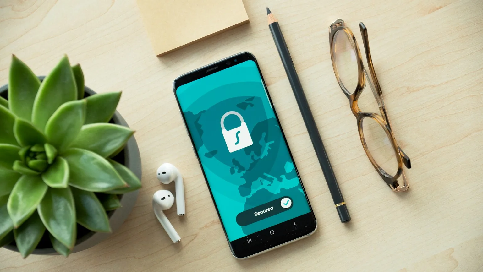 Uno smartphone con l’icona di un lucchetto sullo schermo, su una scrivania con una pianta, degli auricolari e degli occhiali