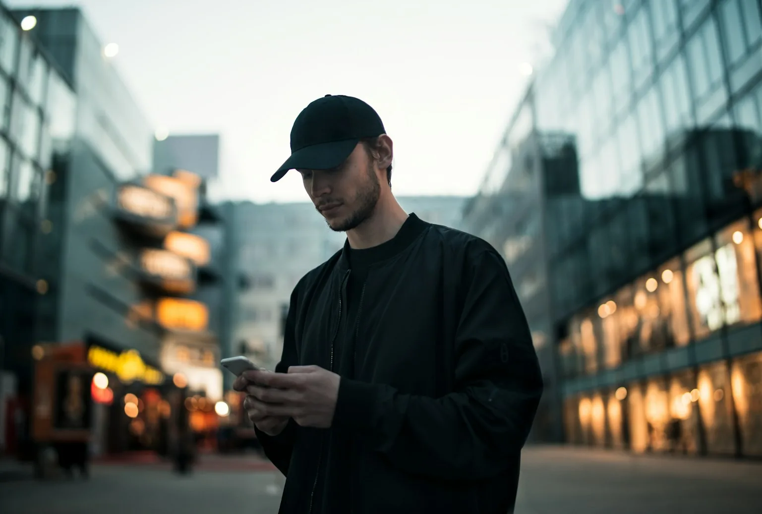 Un ragazzo con un cappellino nero guarda lo smartphone in una strada della città, tra edifici di vetro illuminati