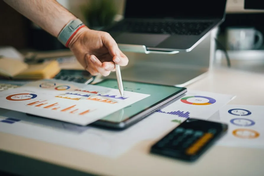 Una mano con una penna indica dei grafici stampati accanto a un tablet e a una calcolatrice