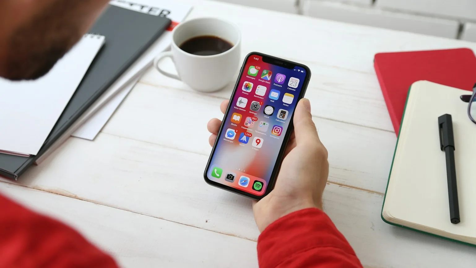 Una persona usa uno smartphone su una scrivania di legno, accanto a una tazza di caffè, a dei quaderni e a una penna