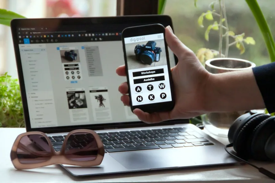Una mano tiene uno smartphone con l’interfaccia di un’app davanti a un portatile che mostra le schermate dello stesso progetto